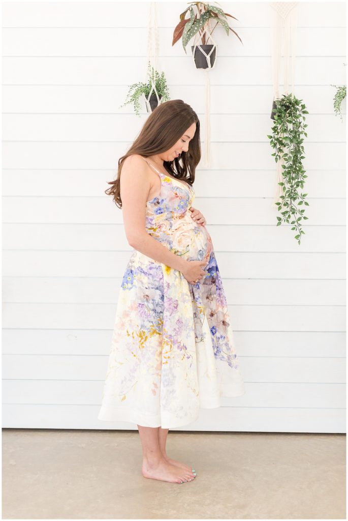 Expecting mother gazes lovingly at pregnant belly while wearing a beautiful floral dress during maternity session with Wisp + Willow Family Photography Team.