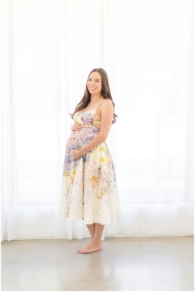 Pregnant woman poses for maternity shoot in McKinney, TX in front of sheer white curtains at Lemon Drop Studio.