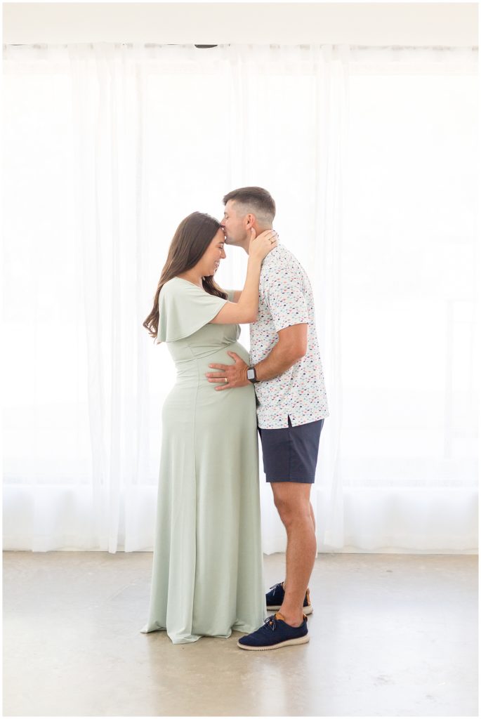 Husband kisses pregnant wife in green dress while holding her expecting belly during maternity session with Wisp + Willow Family Photography Team.