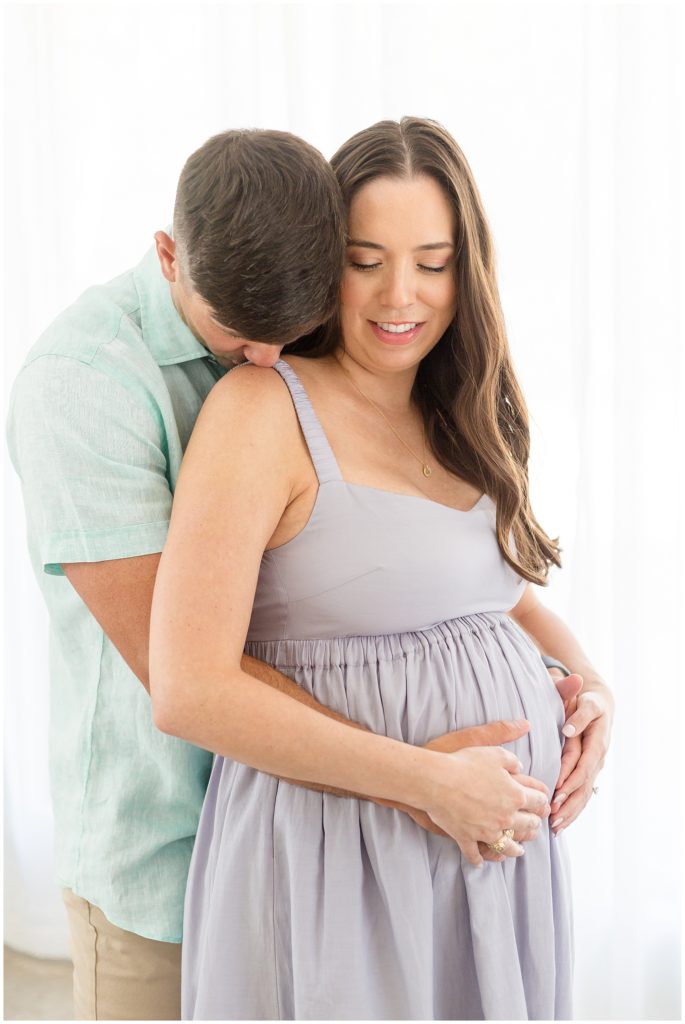 Husband in light blue button down kisses pregnant wife in purple dress on shoulder during Lemon Drop Studio maternity photos.