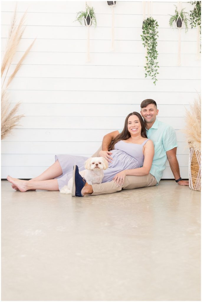 Husband and wife lovingly lounge with white pup during maternity photo session with Wisp + Willow Photography Team.