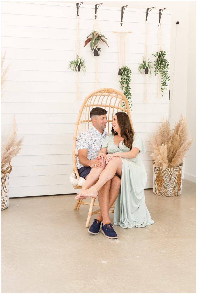 Wife in light green dress sits on husbands lap in wicker chair during maternity studio shoot with Wisp + Willow Photography Team.