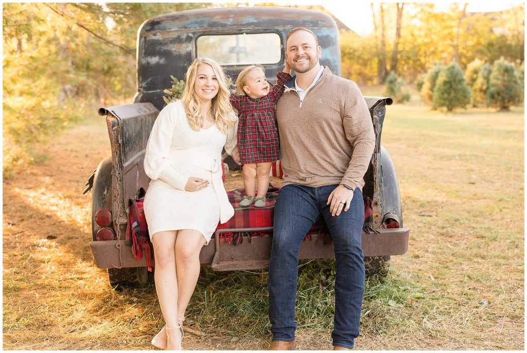 Mom and dad sit in bed of brown pick up truck with toddler daughter in red plaid dress stands in between smiling up at them during Christmas family session. 