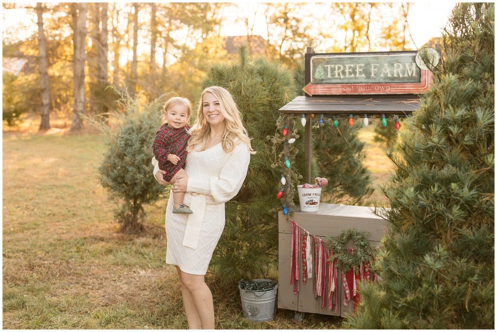 Mom with blonde hair and white sweater dress holds daughter in red and green plaid dress and green shoes during Christmas session. 