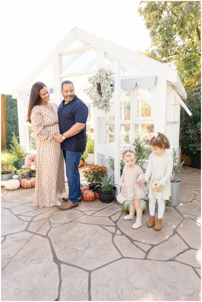 Mom and dad hold hands in front of white greenhouse with 2 daughters holding pumpkins during fall family photo session. 