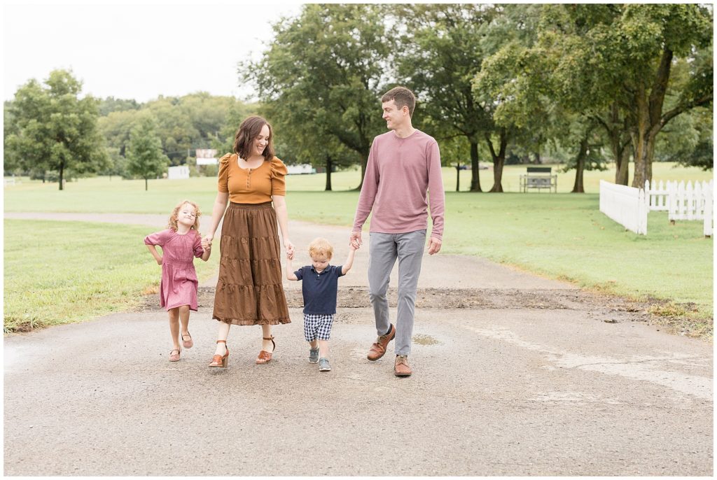 Family of 4 hold hands while walking outside in park during fall family photo session. 