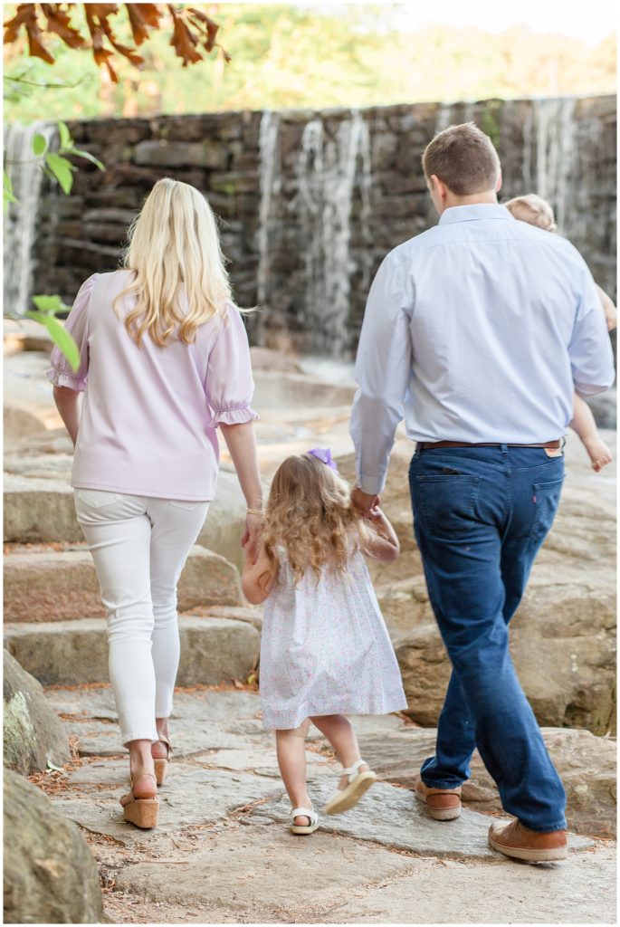 Family of 4 wearing lavender walk while holding hands near beautiful waterfall during summer family photos.