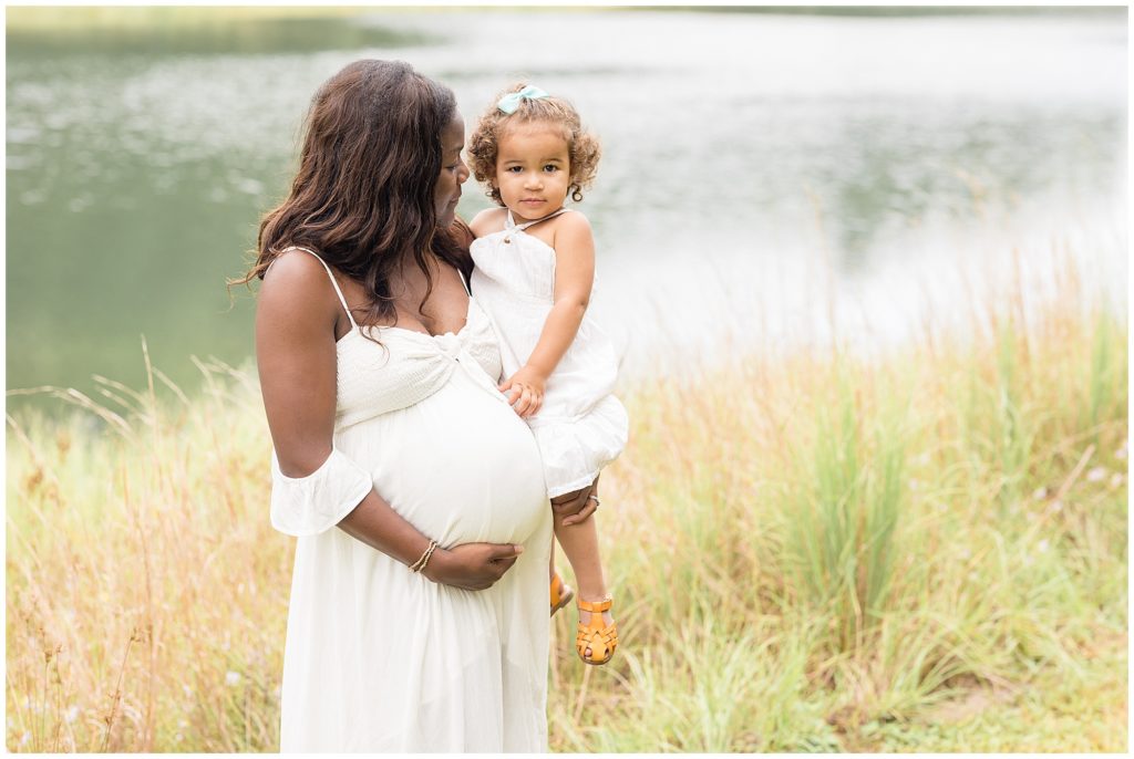 Mom in white dress holds daughter in white dress and bow while standing in long grass with lake behind in Savannah, GA during family maternity session. 