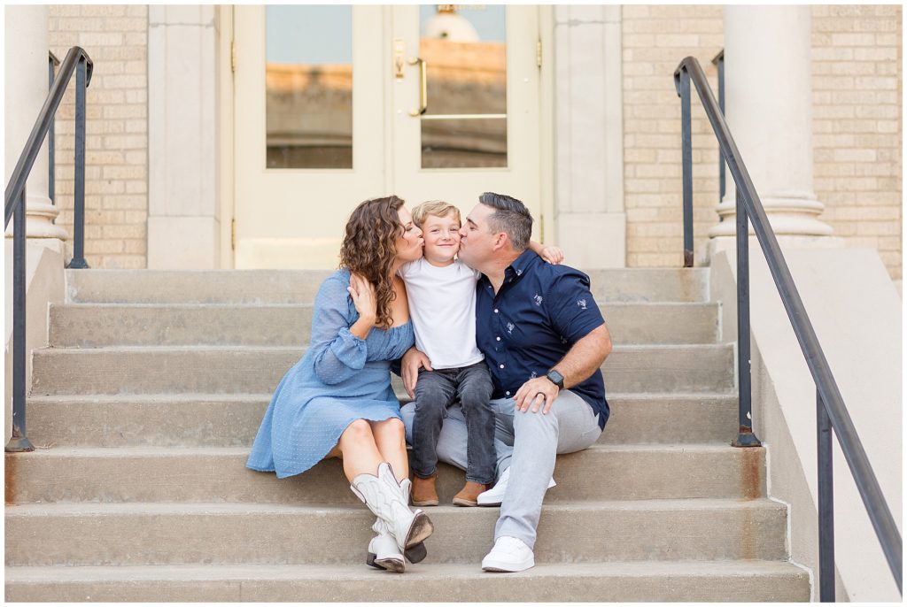 Mom in blue dress and white cowboy boots and dad in dark blue button down and grey pants kiss son on cheeks while sitting on steps in downtown McKinney, TX during family session. 