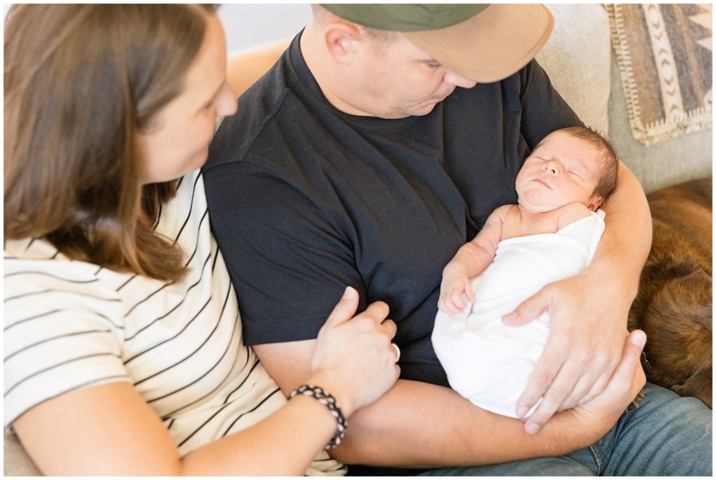 Mom in white stripped shirt and dad in black shirt and hat hold newborn baby in white swaddle while sitting on couch during lifestyle newborn session with Wisp + Willow Photography Co. 