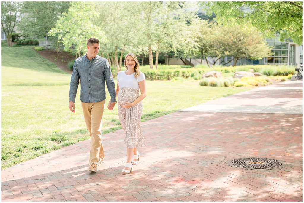 Man in grey button down and woman in tan skirt hold hands and walk down sidewalk with trees in the background during maternity photo session in Richmond, VA. 