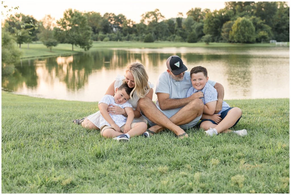 Family of 4 sit in grass in front of pond while playing and giggling together during family session with Wisp + Willow Photography Team in Nashville, TN.