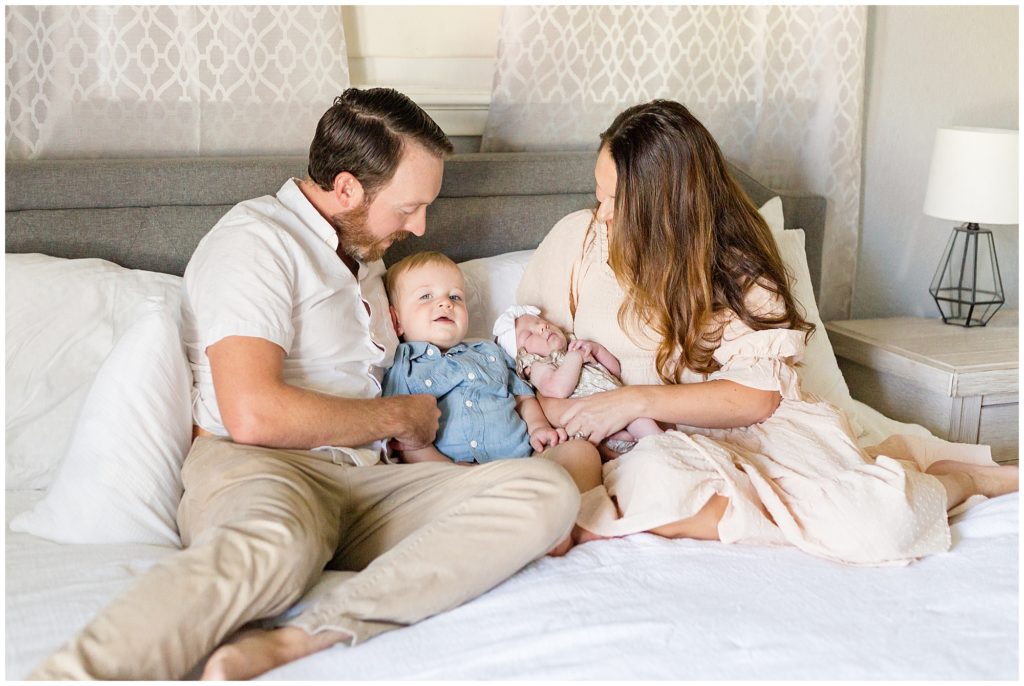 Mom and dad in neutral colors cuddle on white bed with toddler son in blue shirt and newborn baby daughter in white bow during lifestyle newborn session in Dallas, TX. 