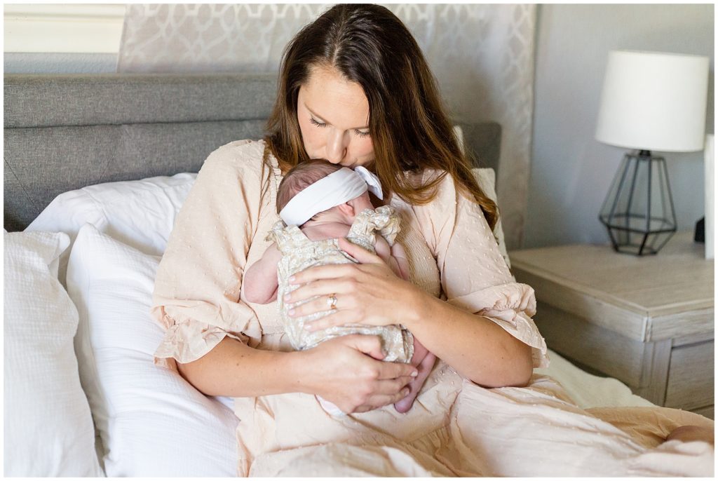 Mom with brown hair and tan dress sits on bed with white linens while holding and kissing her newborn baby daughter in tan and white outfit during newborn session. 