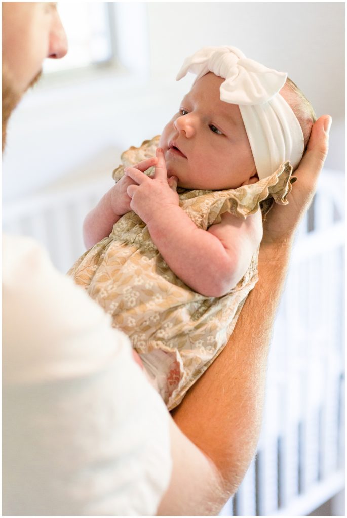 Dad tenderly holds newborn baby daughter in white bow and tan floral outfit during lifestyle newborn session with Wisp + Willow Photography Team. 