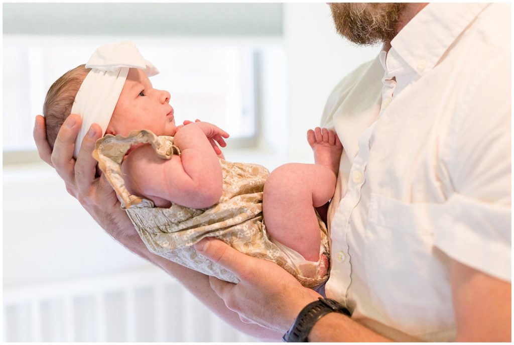 Dad in white button up shirt gently holds newborn baby daughter with white bow and tan ruffle outfit during family session in Dallas, TX. 