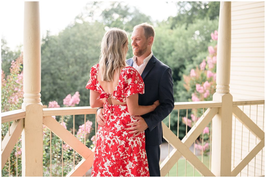 Couple in blue suit and red and white printed dress hold each other on white wooden porch with flowers in the back during photo session. 