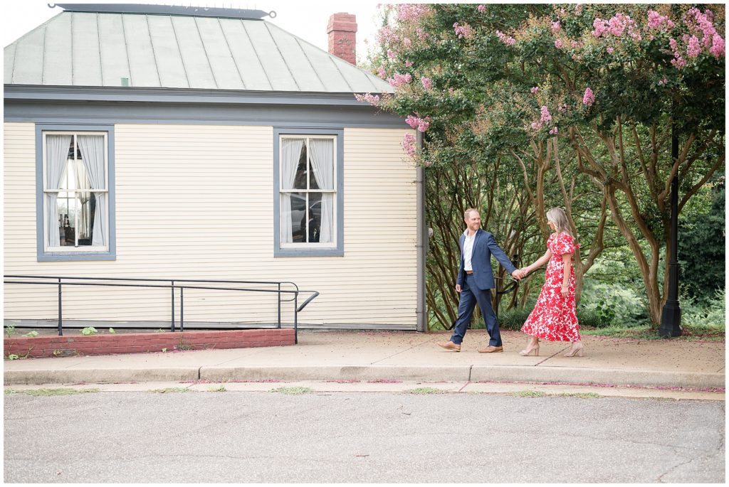 During photo session with Wisp + Willow Photography Co. couple in blue suit and red and white printed dress hold hands and walk in front of history home in Richmond, VA. 
