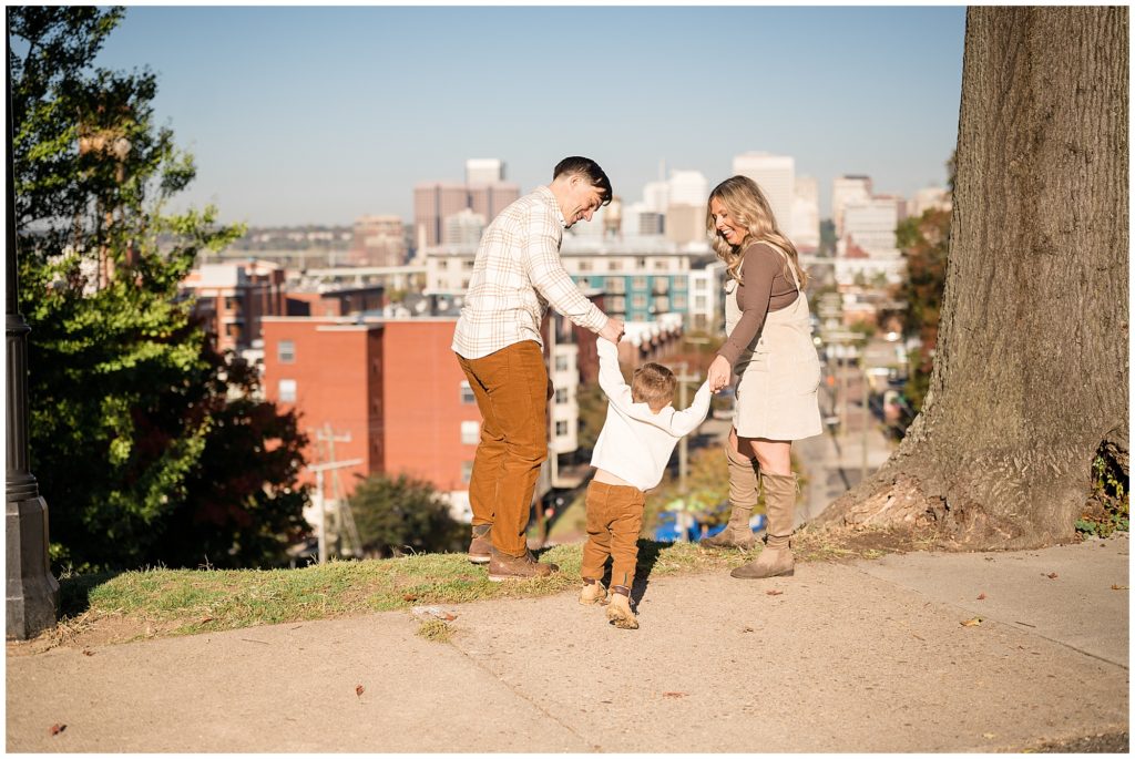 Dad in cream button down and mom in cream jumper hold toddler son's hands while standing in front of the beautiful city skyline of Richmond, VA in Libby Hill. 