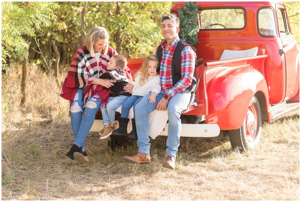 Family of 4 in red plaids sit and snuggle in bed of red pickup truck with Christmas tree in back during family session. 
