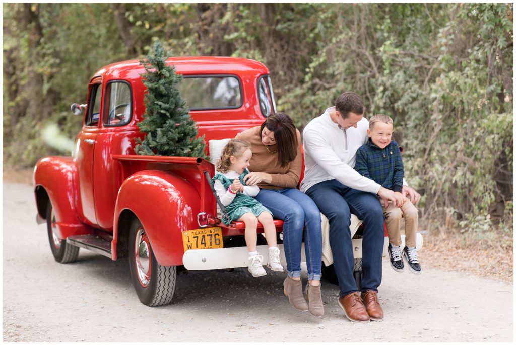 Family of 4 sit and giggle in bed of red pick up truck with pine tree behind them during Christmas family session with Wisp + Willow Photography Co. 