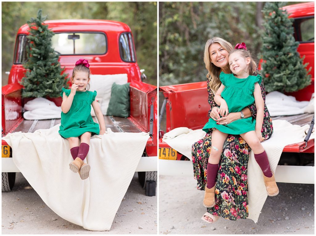 Girl in green dress and green bow sits and smiles on white blanket in bed of red pickup truck. Mom sits in bed of red pickup truck with white blanket and pine tree with daughter in green dress sitting and smiling on her lap during Christmas family session. 