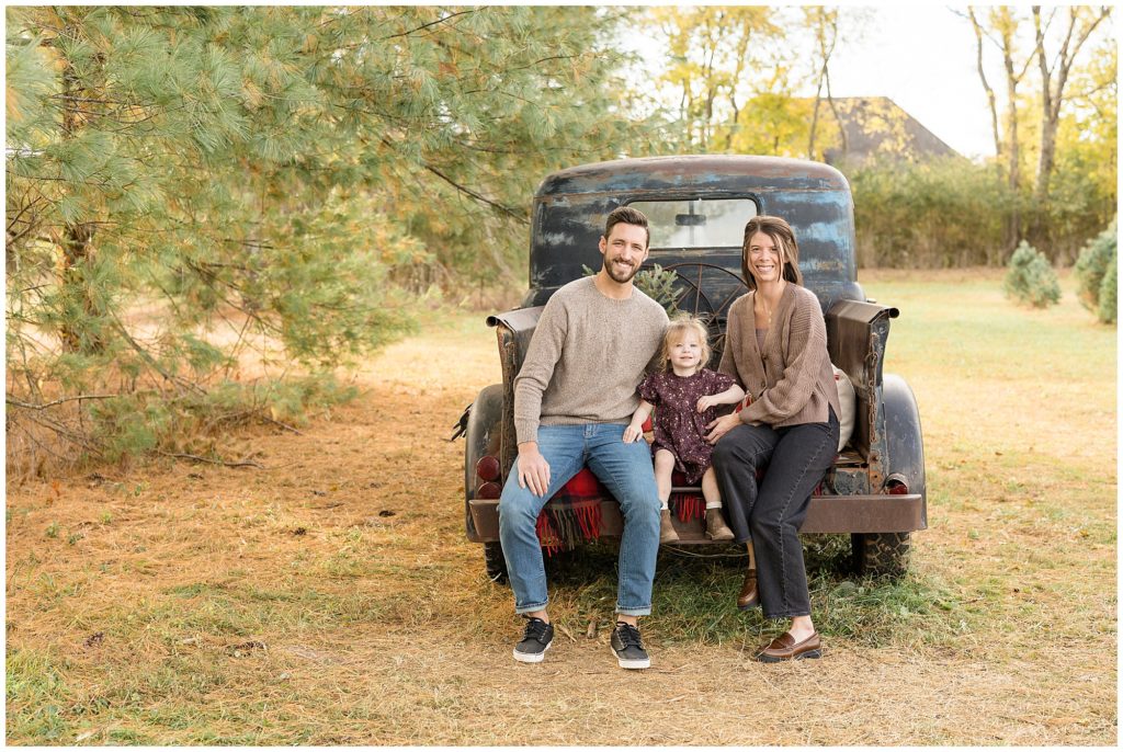 Family of 3 sit in the back of brown pick up truck in field during Christmas photo session with Wisp + Willow Photography Co.