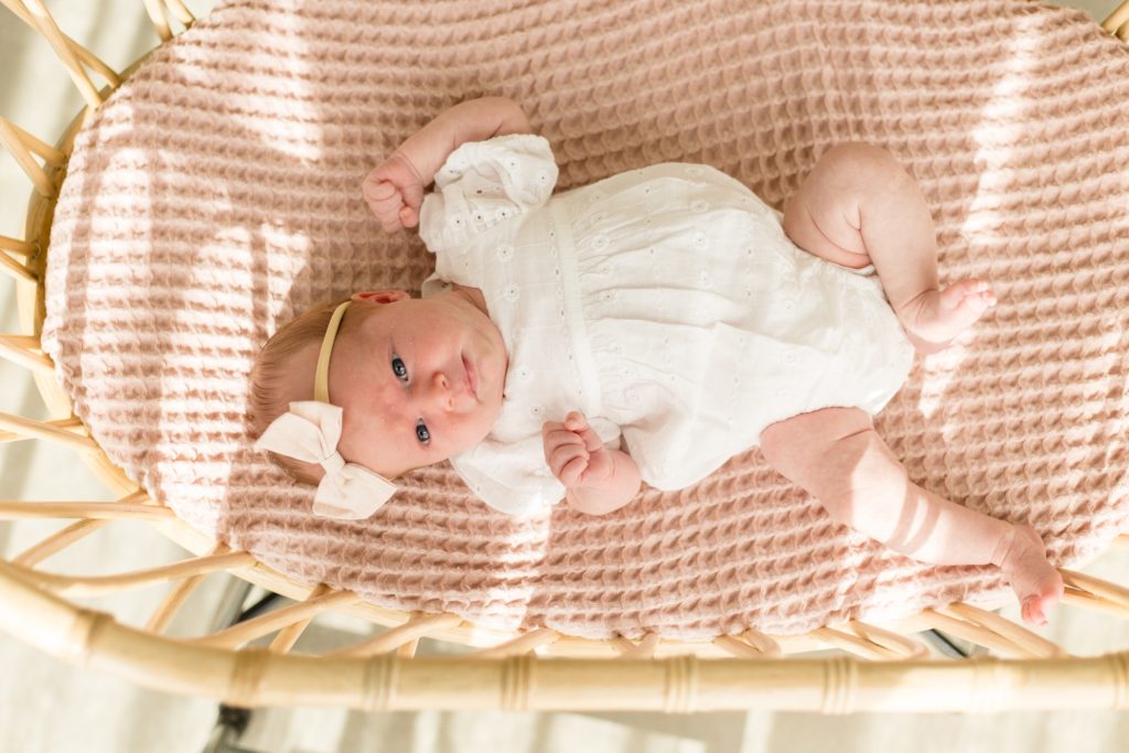 Baby girl in white eyelet onesie lays in bamboo bassinet for lifestyle newborn photos in McKinney, Texas at Lemon Drop Studios with family photography team Wisp + Willow Photography Co. Click to see more from this session live on the blog now! 