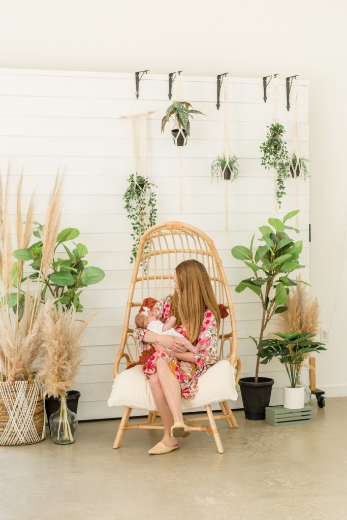 Mom sits in wicker chair in front of white shiplap wall with greenery and wheatgrass during mommy and me lifestyle newborn session with family photography team Wisp + Willow Photography Co. at Lemon Drop Studios in McKinney, Texas. Click to see more from this sweet session live on the blog now! 