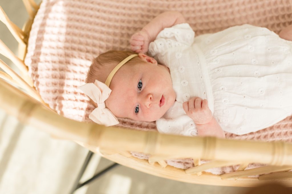 Baby girl in white bow and onesie lays on pink blanket in bamboo bassinet during lifestyle newborn session with family photography team Wisp + Willow Photography Co. in McKinney, Texas at the stunning Lemon Drop Studios 