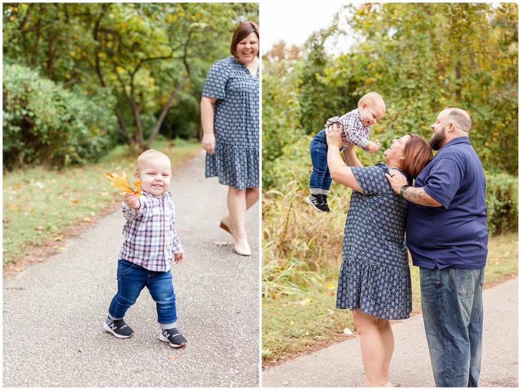 Toddler boy in plaid shirt and jeans walks on path while holding leaf with mom in the background. Mom in blue dress and dad in blue shirt hold son up in the air during fall family photo session in Raleigh, NC.
