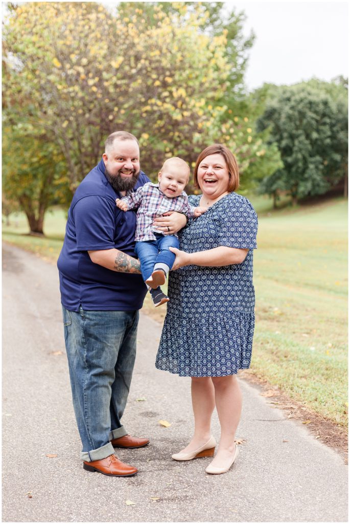 Mom and dad in blue dress and shirt hold baby son in plaid shirt while laughing in Dorothea Dix Park during fall family photo session.