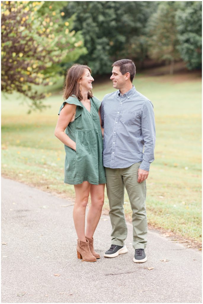 Husband in blue plaid button down and wife in green dress stand together and look in each other eyes during fall family photo session outside at Dorothea Dix Park. 