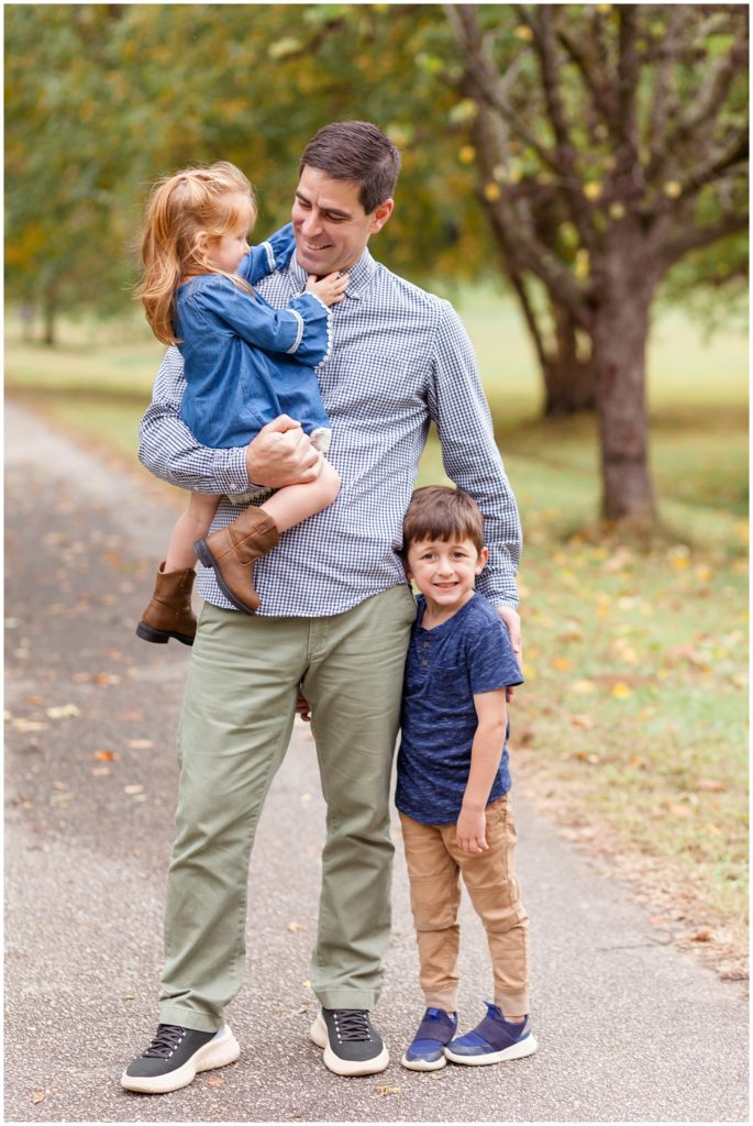 Dad holds daughter in blue denim dress and brown boots while son in blue shirt and brown pants hugs his leg during fall family photo session in Dorothea Dix Park. 