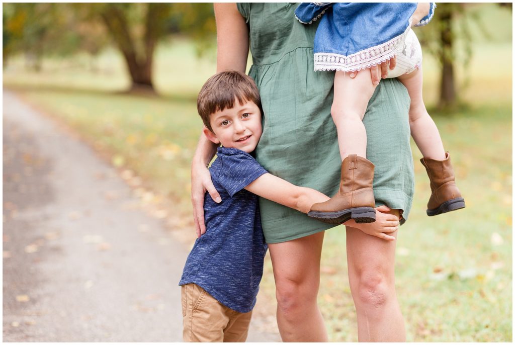 Little boy in dark blue shirt hugs onto mom's legs while mom holds little girl in brown boots during fall family session in Raleigh, NC.