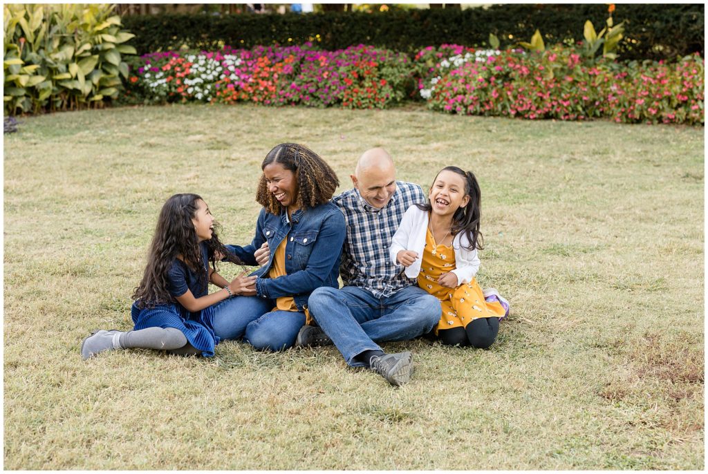 Mom and Dad and 2 daughters sit in grassy field with flowers behind them laughing and playing during outdoor family photo session with Wisp + Willow Photography Co.