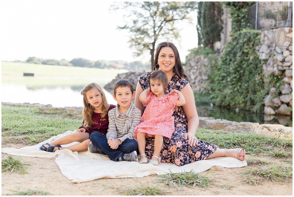 Mom and 3 kids sit and pose on white blanket in front of rock wall and pond during outdoor family photo session with Wisp + Willow Photography Co.  