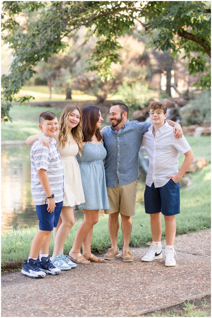 Mom, dad, and 3 teenagers stand with their arms around each other on sidewalk in front of pond in Frisco Central Park in Frisco, TX. 