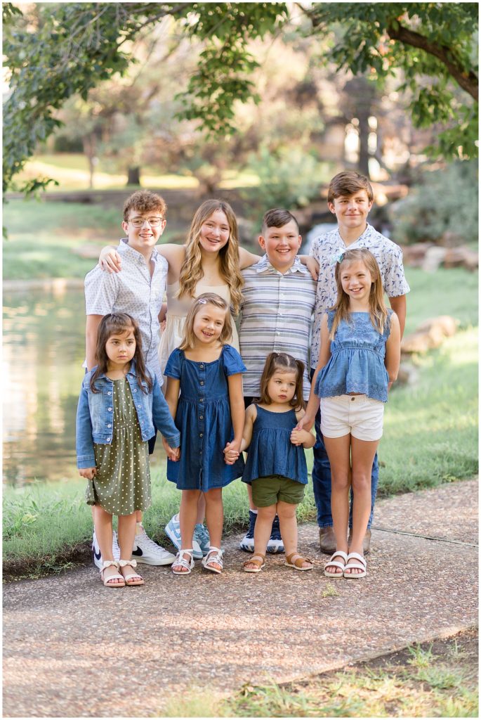 8 kids in different shades of blue stand with arms around each other smiling in front of pond outside at Frisco Central Park in Texas during extended family session. 