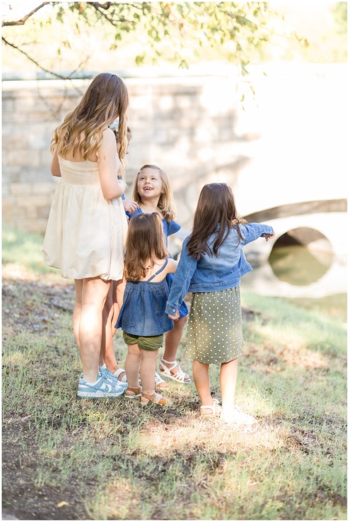 Group of girls play and smile outside in Frisco Central Park in front of pond and brick wall during family photography session with Wisp + Willow Family Photography Team. 
