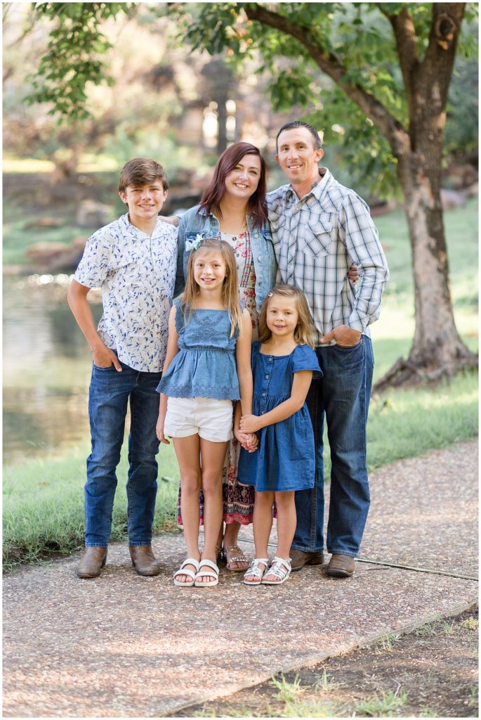Family of 5 in different shades of blue stand and pose in front of lake outside in Frisco, TX during family photo session with Wisp + Willow Photography Co.