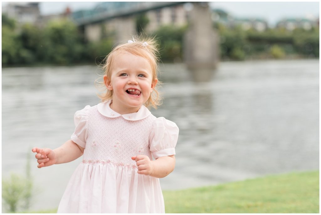Little girl in pale pink dress giggles and plays in front of Tennessee River in Chattanooga, TN during family maternity session with Wisp + Willow Family Photography Team. 