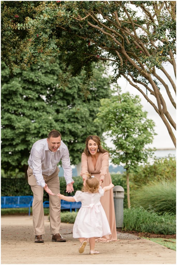 Dad and mom reach out to daughter in pale pink dress and bow while she runs into their arms on sidewalk under trees in Coolidge Park during family maternity session. 