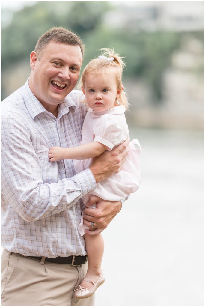 Dad in plaid button down and tan khakis holds daughter in pale pink dress and bow during family maternity session with Wisp + Willow Family Photography Team. 