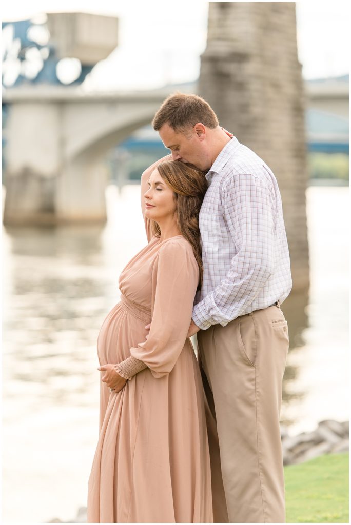 Expectant wife in blush dress leans on husband in plaid button down and tan pants while he kisses her head in front of the Tennessee River during family maternity session in Chattanooga, TN. 
