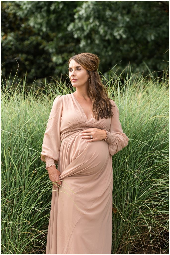 Expecting woman in flowy blush dress poses in front of tall grass in Coolidge Park in Chattanooga, TN during maternity session with Wisp + Willow Family Photography Team. 