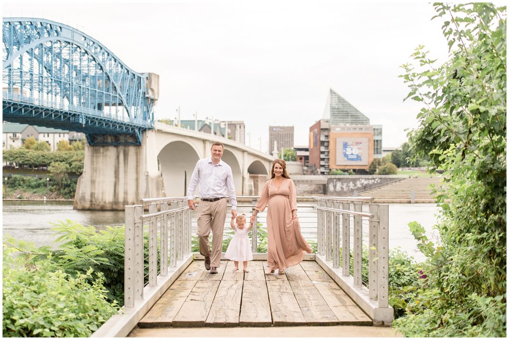 Family of 3 hold hands and walk down pier on Tennessee River in Chattanooga, TN during family maternity session with Wisp + Willow Photography Team. 