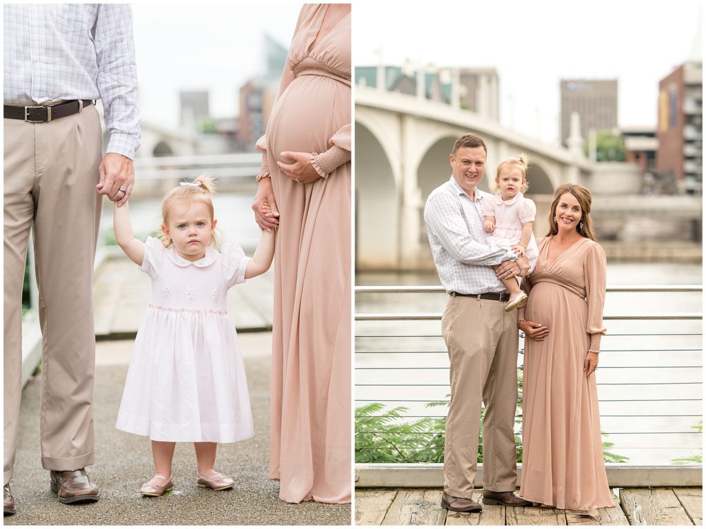 Mom and dad hold toddler daughter's hands on bridge in Coolidge Park during family maternity session. Dad holds daughter in pale pink dress while mom in blush dress places her hand on her belly in front of the Tennessee River. 