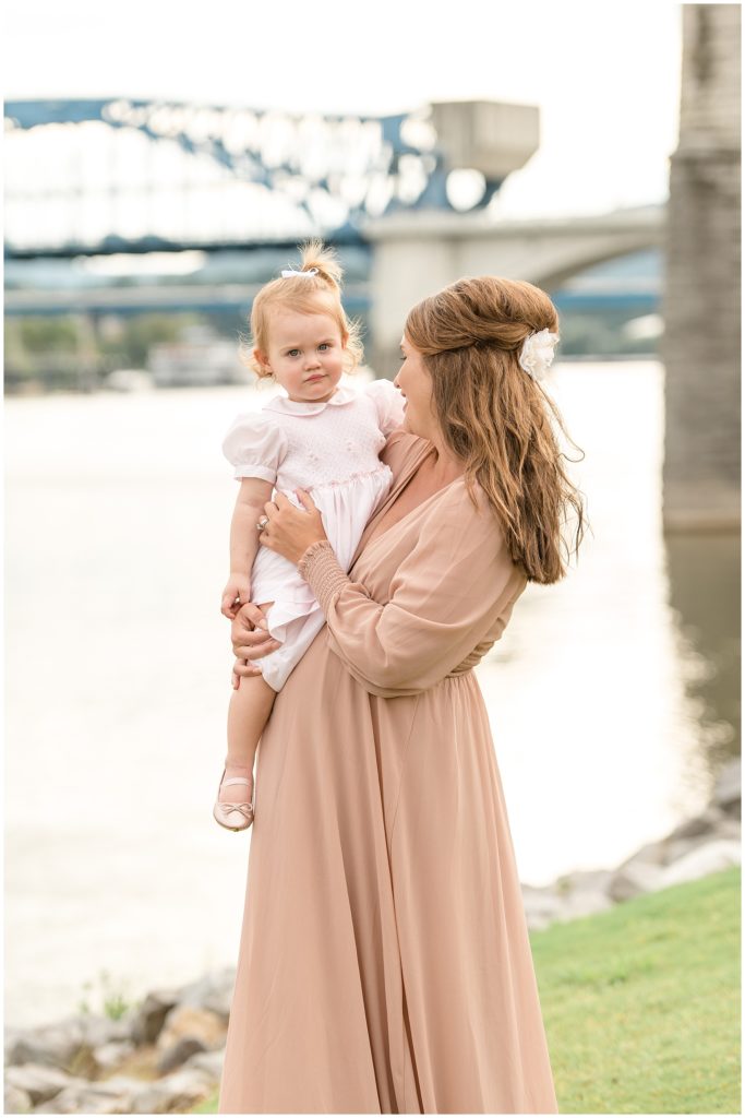Expecting mother in blush dress and white flower in hair holds daughter in pale pink dress in front of Tennessee River in Coolidge Park in Chattanooga, TN. 