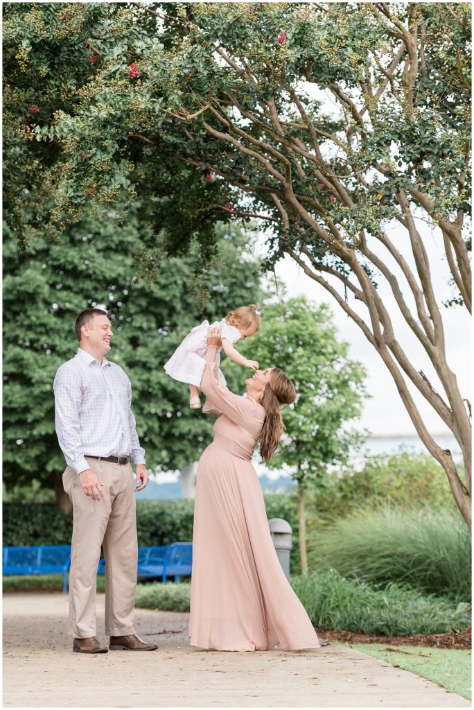 Pregnant mom in blush dress holds toddler daughter in pale pink dress overhead while dad smiles at them underneath beautiful trees in Coolidge Park. 